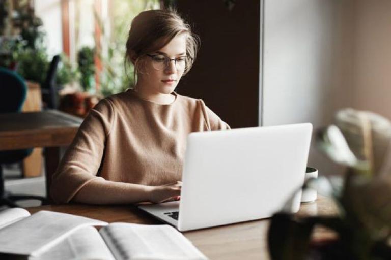 Woman working on her laptop, possibly at home; some books on the table and some plants in the background
